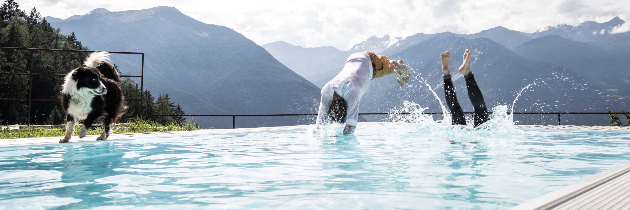 Pool mit Bergpanorama, zwei Personen beim Sprung ins Wasser und Hund am Beckenrand
